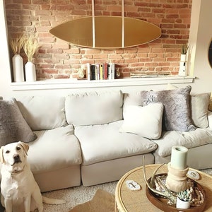 K&ouml;nnte beinhalten: Ein wei&szlig;es Ecksofa mit einem hellgrauen Kissen und einem flauschigen grauen Kissen. Ein wei&szlig;er Hund sitzt vor dem Sofa. Ein Couchtisch aus Holz mit einer geflochtenen Tischplatte hat eine Holzschale mit Geweih, eine kleine Topfpflanze und eine Kerze.