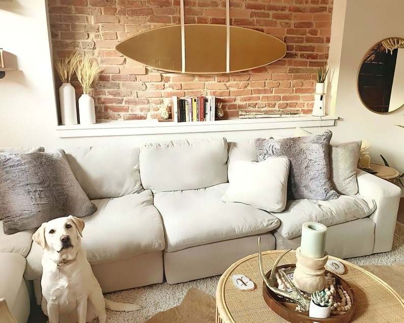 K&ouml;nnte beinhalten: Ein wei&szlig;es Ecksofa mit einem hellgrauen Kissen und einem flauschigen grauen Kissen. Ein wei&szlig;er Hund sitzt vor dem Sofa. Ein Couchtisch aus Holz mit einer geflochtenen Tischplatte hat eine Holzschale mit Geweih, eine kleine Topfpflanze und eine Kerze.