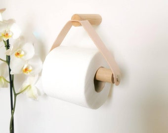 May include: A wooden toilet paper holder with a light brown leather strap. The holder is mounted to a white wall and has a roll of white toilet paper on it.