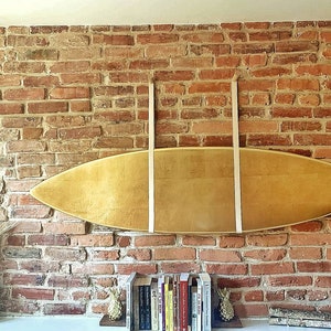 K&ouml;nnte beinhalten: Ein h&ouml;lzernes Surfbrett h&auml;ngt an einer Backsteinwand mit zwei wei&szlig;en Lederriemen. Das Surfbrett ist hellbraun und hat eine nat&uuml;rliche Holzmaserung. Unter dem Surfbrett befinden sich B&uuml;cher und zwei Ananas-Dekorationen auf einem Regal.