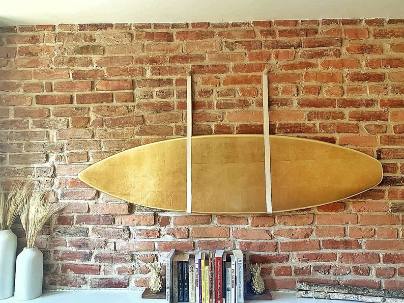 K&ouml;nnte beinhalten: Ein h&ouml;lzernes Surfbrett h&auml;ngt an einer Backsteinwand mit zwei wei&szlig;en Lederriemen. Das Surfbrett ist hellbraun und hat eine nat&uuml;rliche Holzmaserung. Unter dem Surfbrett befinden sich B&uuml;cher und zwei Ananas-Dekorationen auf einem Regal.
