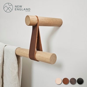 Puede incluir: Barra de toalla de madera con una correa de cuero. La barra est&aacute; montada en una pared blanca. La correa es marr&oacute;n y tiene una forma triangular. La barra est&aacute; hecha de madera natural. La barra de toalla es parte de un conjunto de accesorios de ba&ntilde;o.