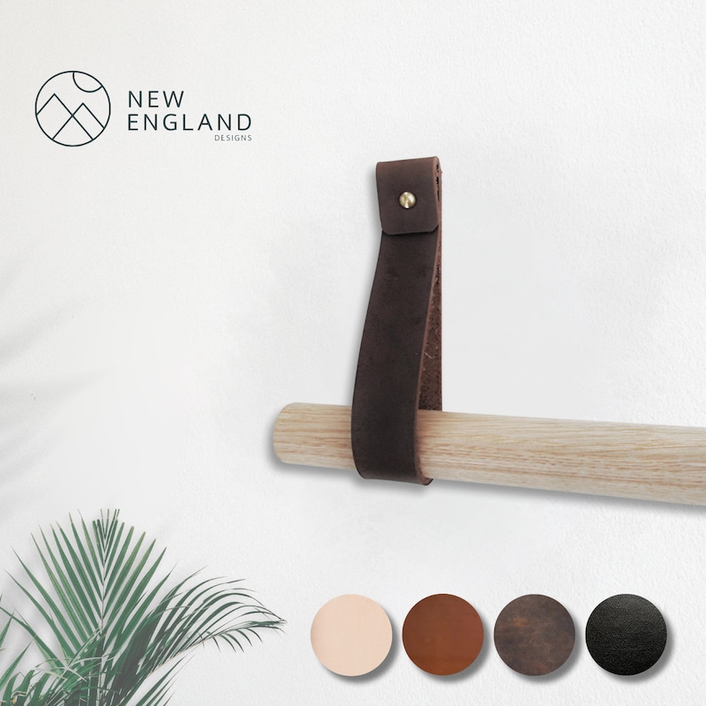 May include: A brown leather towel bar hanger with a brass screw. The hanger is attached to a wooden dowel rod. The background is a white wall.  New England Designs.