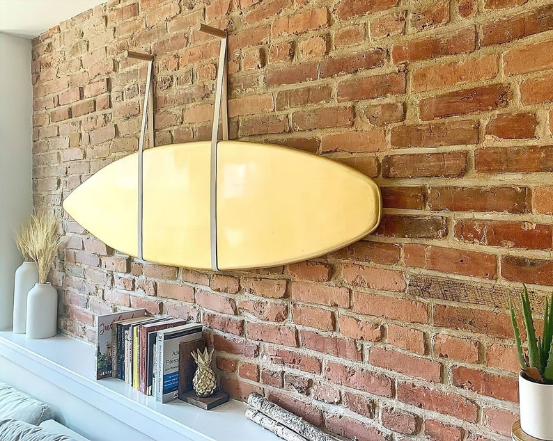 K&ouml;nnte beinhalten: Ein gelbes Surfbrett h&auml;ngt an einer Backsteinwand mit zwei Lederriemen. Das Surfbrett h&auml;ngt &uuml;ber einem Regal mit B&uuml;chern und einer Ananasfigur.