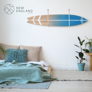 K&ouml;nnte beinhalten: Ein blaugr&uuml;nes und wei&szlig;es Surfbrett mit Holzmaserung h&auml;ngt an einer wei&szlig;en Wand &uuml;ber einem Bett mit einer blaugr&uuml;nen Decke und Kissen. Das Surfbrett hat an jedem Ende zwei wei&szlig;e Pfeile. Der Text "NEW ENGLAND DESIGNS" ist in der oberen linken Ecke des Bildes sichtbar.