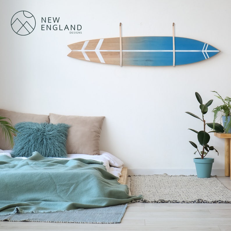 K&ouml;nnte beinhalten: Ein blaugr&uuml;nes und wei&szlig;es Surfbrett mit Holzmaserung h&auml;ngt an einer wei&szlig;en Wand &uuml;ber einem Bett mit einer blaugr&uuml;nen Decke und Kissen. Das Surfbrett hat an jedem Ende zwei wei&szlig;e Pfeile. Der Text "NEW ENGLAND DESIGNS" ist in der oberen linken Ecke des Bildes sichtbar.