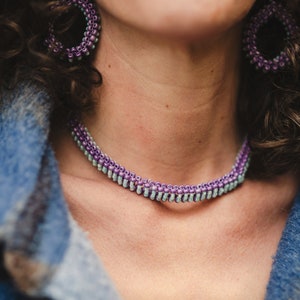 May include: A close-up of a woman's neck wearing a purple and light blue beaded choker necklace. The necklace is made of small, round beads that are strung together in a continuous pattern. The woman is also wearing matching purple and light blue beaded earrings.