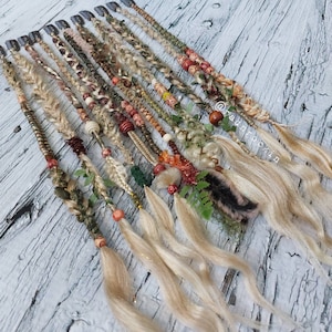 May include: A selection of handcrafted hair extensions featuring diverse textures and decorations. The braids are embellished with beads, leaves, and feathers in earthy tones of brown, beige, and green. Each extension has a metal clip for attachment.