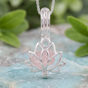May include: A silver-toned metal pendant necklace featuring a cage-style locket shaped like a lotus flower. The locket contains a single, raw, pale pink gemstone inside its openwork frame. The pendant hangs from a delicate silver-toned chain.