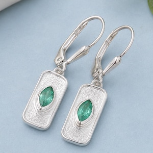 May include: A pair of silver drop earrings featuring a rectangular pendant with a textured, matte finish. Each pendant is centered with a single marquise-cut green gemstone held in a bezel setting, attached to a secure silver leverback closure.