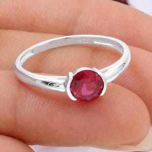 May include: A silver ring with a round, deep red gemstone. The ring band is smooth and polished, with the marking "925" visible. The gemstone is set in a simple, classic design, showcasing its vibrant color.