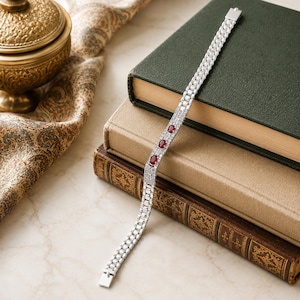 May include: A silver bracelet with three red gemstones and multiple small white stones. The bracelet is displayed on a marble surface next to a stack of books and a decorative gold container.