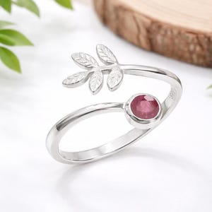 May include: A silver ring with a leaf design and a round, red gemstone. The ring features a delicate leaf motif on one end and a bezel-set, ruby-colored stone on the other. The ring is set against a soft, white background.