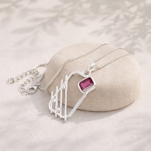 May include: A silver necklace with a geometric pendant featuring a rectangular, magenta gemstone. The pendant has a unique design with vertical bars and small, round accents. The chain is delicate and adjustable.