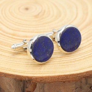 May include: A pair of cufflinks with a round, deep blue stone set in a silver-colored metal. The stone has flecks of gold throughout. The cufflinks are displayed on a light brown wooden surface.