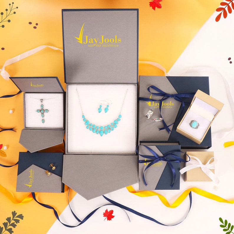 K&ouml;nnte beinhalten: Eine Auswahl an Schmuckboxen in verschiedenen Gr&ouml;&szlig;en und Farben mit blauen und goldenen Schleifenakzenten. Die Boxen sind ge&ouml;ffnet und zeigen t&uuml;rkisfarbenen und silbernen Schmuck, darunter eine Halskette, Ohrringe, einen Ring und einen Kreuzanh&auml;nger. Die Boxen sind mit dem Markennamen "Jay Jools, feel the excellence." beschriftet.