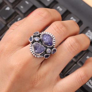 May include: A silver ring featuring multiple irregularly shaped, deep blue gemstones. The ring has a textured, beaded border and is set with various sizes of blue and purple stones, creating a unique and eye-catching design.