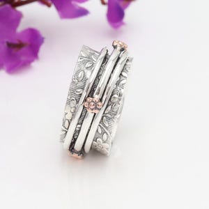May include: A silver spinning ring with a floral design and rose gold accents. The ring is made of multiple bands that spin freely.