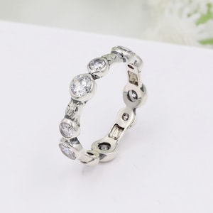 May include: A silver-toned ring featuring a series of clear, round gemstones set in a repeating pattern. The ring has a detailed, textured band with leaf-like designs. The gemstones are evenly spaced around the ring's circumference.