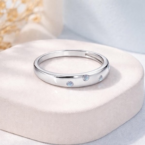 May include: A silver ring with a polished finish. The ring features small, round, light blue gemstones evenly spaced around the band. The ring is displayed on a soft, light-colored surface.