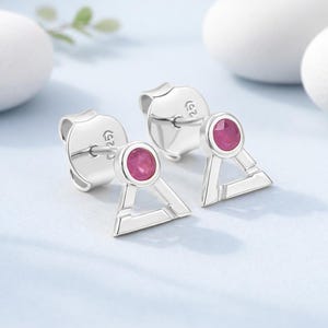 May include: A pair of silver stud earrings with a geometric triangle design. Each earring features a small, round, pink gemstone set in a bezel setting. The earrings have a post and butterfly backing.