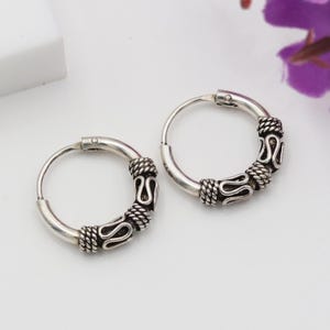 May include: A pair of silver hoop earrings. Each earring has a decorative section with a rope-like texture and a stylized design. The earrings are on a white surface, with a purple flower in the background.