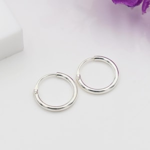 May include: Two silver hoop earrings. The earrings are circular and have a smooth, shiny finish. They are displayed on a white surface, with a hint of purple flowers in the background. The earrings are a classic jewelry item.