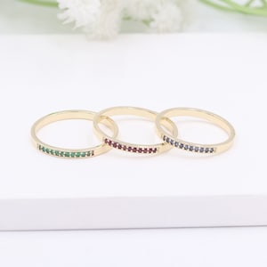 May include: Three gold rings, each set with a row of small gemstones. One ring features green stones, another red, and the third blue. The rings are presented on a white surface.