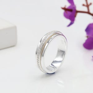 May include: A silver ring featuring a row of small, clear gemstones. The ring has a double-band design, with one band overlapping the other. The ring is set against a white background with a purple flower in the background.