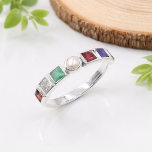 May include: A silver ring featuring a central pearl and a series of colorful gemstones. The gemstones include square-cut emerald, ruby, sapphire, and a clear stone, set in a band. The ring is displayed on a white surface.