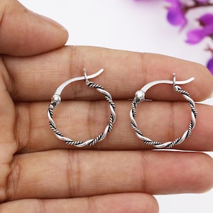 May include: A pair of silver hoop earrings with a twisted rope design. The earrings are round and have a hinged closure. The earrings are held in a hand against a soft background.