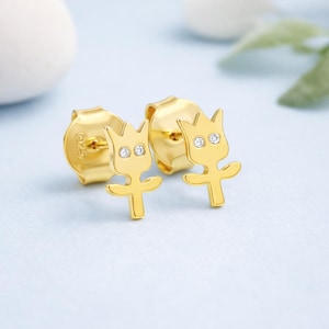 May include: A pair of gold-colored stud earrings. Each earring features a stylized flower design with a crown-like top and outstretched arms. The flower's center has two small, sparkling, clear gemstones. The earrings are on a light blue background.