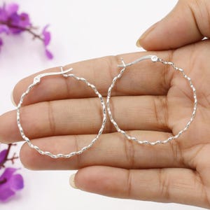 May include: A pair of silver hoop earrings with a twisted rope design. The earrings are round and have a simple clasp closure. The earrings are held in a person's hand.