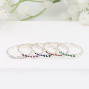 May include: Five silver stacking rings with a single row of colorful gemstones. The rings are arranged in a row, with the white gemstone ring on the left and the green gemstone ring on the right. The rings are all the same size and have a simple, elegant design.