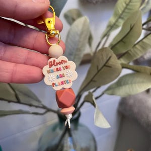 Puede incluir: Un llavero de color rosa y blanco con forma de flor con el texto "Bloom where you are planted" en letras de colores. El llavero tiene un anillo de llave dorado y una cadena de cuentas rosas y blancas.
