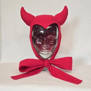 May include: A red devil costume hood with curved horns and a large bow. The hood is made of a soft fabric and is designed to be worn on the head. The horns are also made of the same red fabric and are attached to the top of the hood.