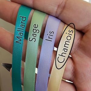 May include: Four spools of ribbon in different colors: teal, sage green, lavender, and cream. The ribbon spools are labeled "Mallard", "Sage", "Iris", and "Chamois".