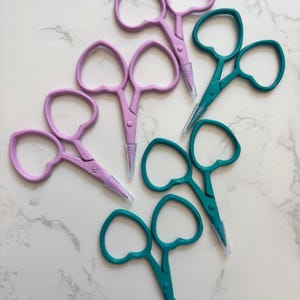 May include: An overhead shot displays several pairs of small scissors. The scissors have heart-shaped finger holes and come in shades of teal and lavender. Each pair has a clear plastic tip cover. The scissors are arranged on a white marble surface.