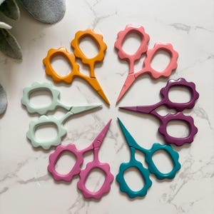 May include: A collection of six pairs of small scissors arranged in a circular pattern. Each pair features a distinct solid color including orange, light pink, purple, teal, magenta, and mint green, with scalloped, flower-shaped finger loops.