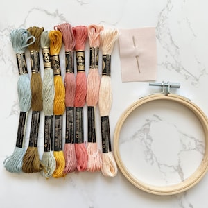 May include: A set of embroidery floss in shades of blue, green, yellow, pink, and beige, along with a needle, a piece of fabric, and a wooden embroidery hoop.