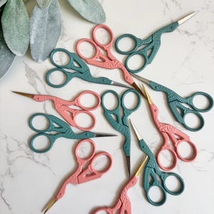 May include: A collection of stork-shaped embroidery scissors in matte pink and muted teal colors. Each pair of scissors features a bird-like design with pointed metal blades, circular finger loops, and textured handles, arranged in a scattered pattern.