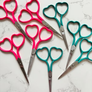 Puede incluir: Un juego de seis tijeras en forma de corazón, tres rosas y tres turquesa, con cuchillas plateadas. Las tijeras están dispuestas sobre una superficie de mármol blanco.