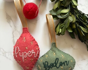 Könnte beinhalten: Zwei Filz-Weihnachtsbaumschmuck mit goldfarbenem Glitzerband. Die Anhänger sind tropfenförmig und haben bestickte Namen: "Piper" und "Boden".