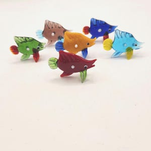 May include: A collection of six colorful glass fish figurines. Each fish features a unique color scheme, including green, brown, orange, blue, red, and light blue. The fish have detailed fins and tails, and are arranged on a white surface.
