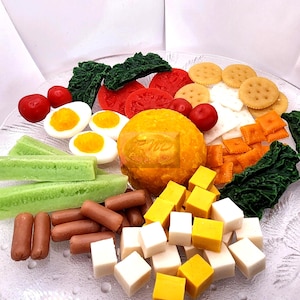 May include: A clear glass plate displays a colorful assortment of miniature food items. The arrangement includes red tomatoes, green cucumbers, yellow cheese cubes, and brown sausages. There are also crackers, eggs, and a large orange item.