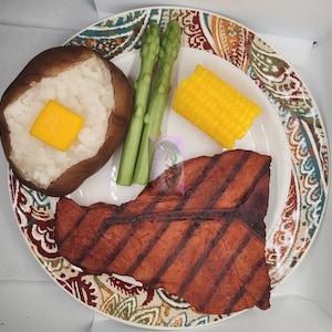 May include: A plate with a brown and white baked potato, a yellow corn cob, green asparagus, and a grilled steak. The plate has a colorful paisley pattern.