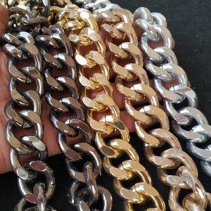 以下が含まれることがあります： さまざまな金属仕上げの太いチェーンネックレスのコレクションを手に持っています。チェーンはシルバー、ゴールド、ガンメタルの色合いで、大胆でモダンなジュエリースタイルを表現しています。リンクは大きく丸みを帯びています。