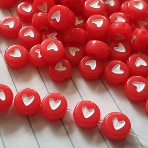 May include: A pile of red heart-shaped beads with white hearts in the centre. The beads are scattered on a white and red striped surface.