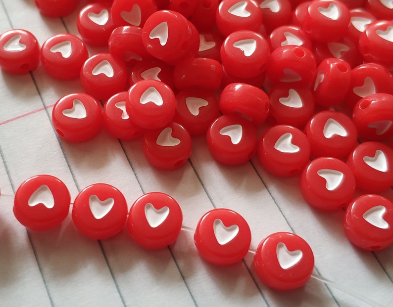 May include: A pile of red heart-shaped beads with white hearts in the centre. The beads are scattered on a white and red striped surface.