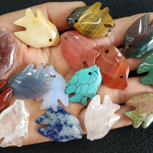 May include: A collection of gemstone fish pendants in various colors, including red, green, blue, and yellow. Each pendant is carved with a unique pattern and has a hole for stringing. The pendants are held in a hand.